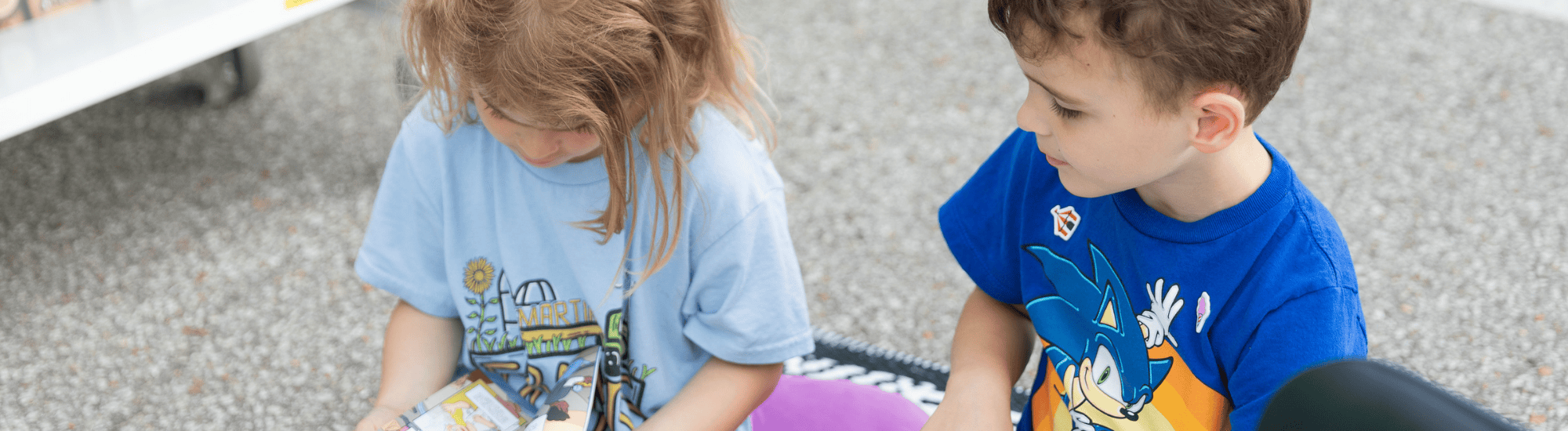 Two children are sitting and reading books on the ground.