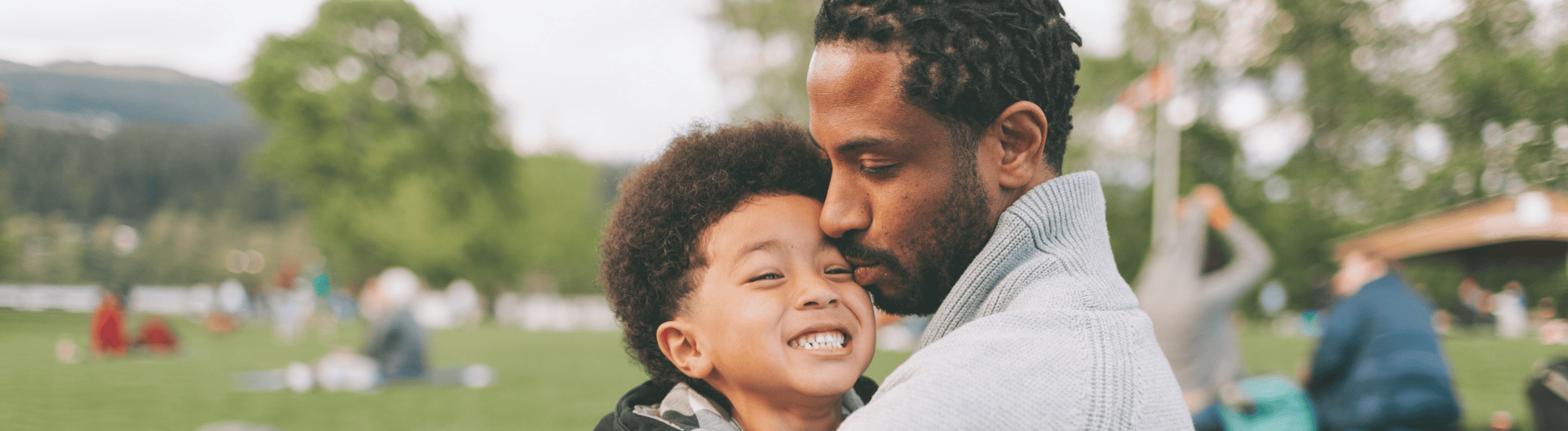 A boy is sitting with his father while being kissed by him.